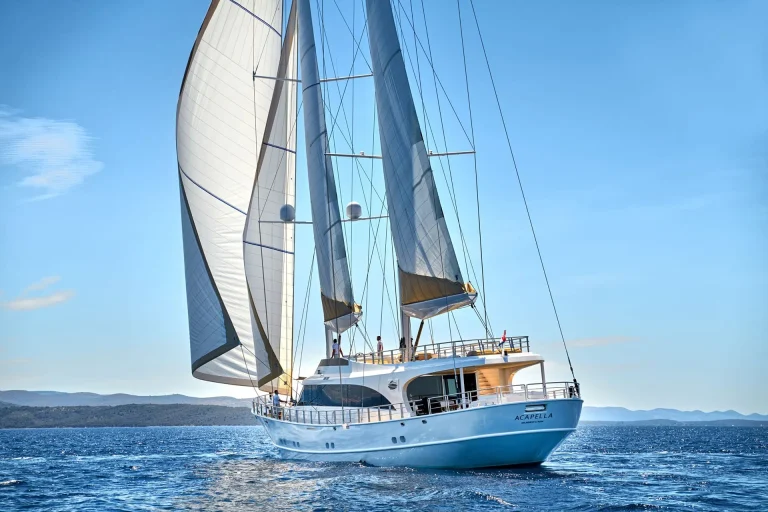 A large white sailboat named Acapella sails on calm blue water under clear skies, with its sails fully unfurled. The coastline and distant hills are visible in the background.