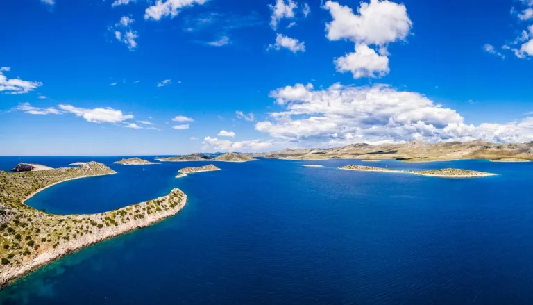 Aerial view of small islands with rocky shorelines and sparse vegetation in a bright blue sea under a partly cloudy sky. Rugged hills are visible in the background.