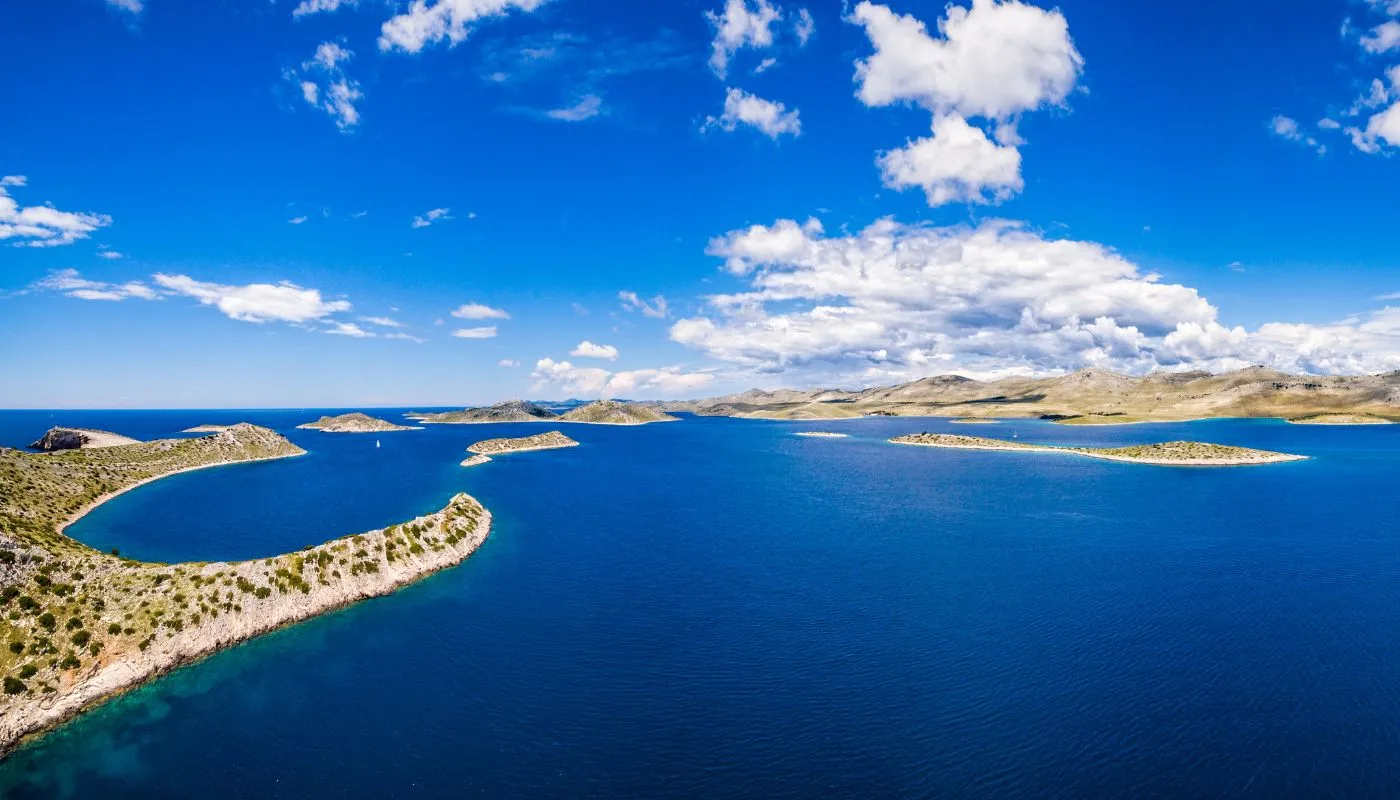 Aerial view of small islands with rocky shorelines and sparse vegetation in a bright blue sea under a partly cloudy sky. Rugged hills are visible in the background.