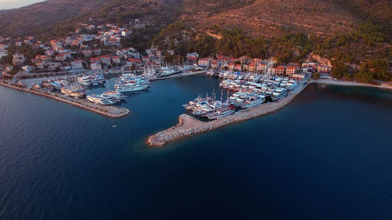 Aerial view of a marina with numerous yachts and boats docked, surrounded by a coastal town with red-roofed buildings and green hills, next to deep blue water at sunset.