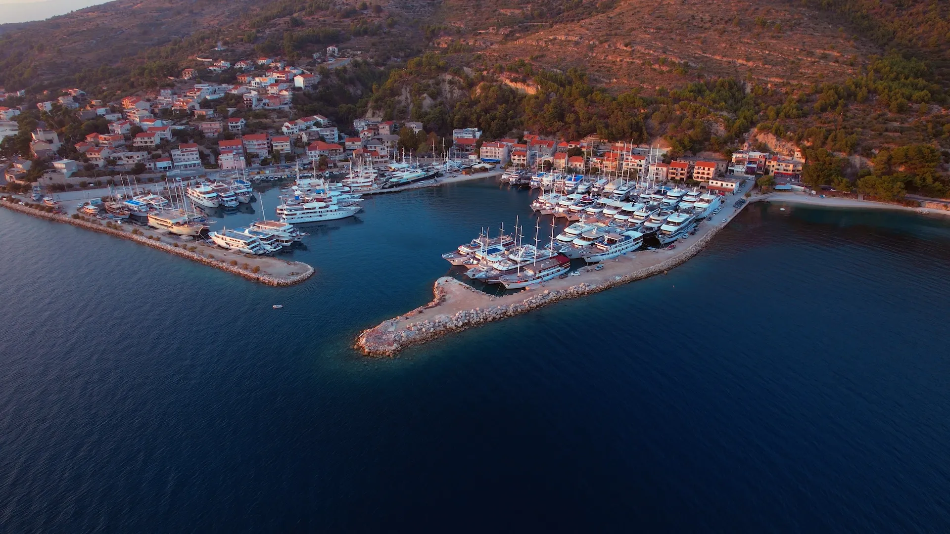 Aerial view of a marina with numerous yachts and boats docked, surrounded by a coastal town with red-roofed buildings and green hills, next to deep blue water at sunset.