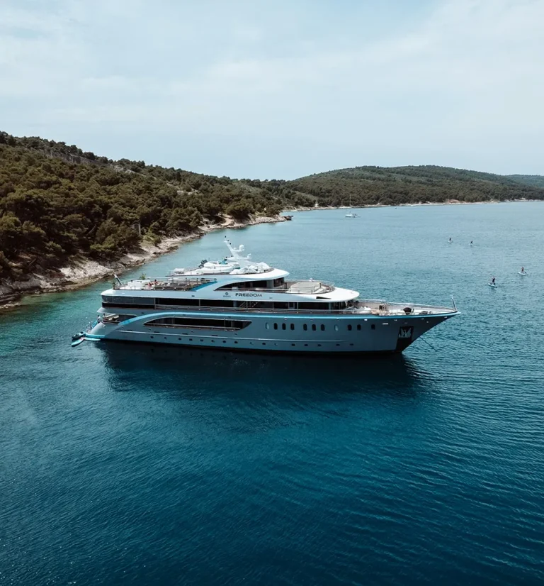 A large white yacht is anchored in calm blue water near a forested coastline with hills in the background under a partly cloudy sky. Paddleboarders are visible in the distance.