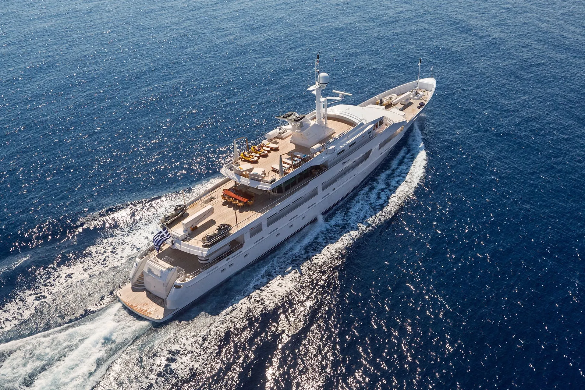 A large, luxurious yacht sails through deep blue ocean water, leaving white waves in its wake. The vessel has multiple decks with seating areas and sun loungers, and is photographed from above.