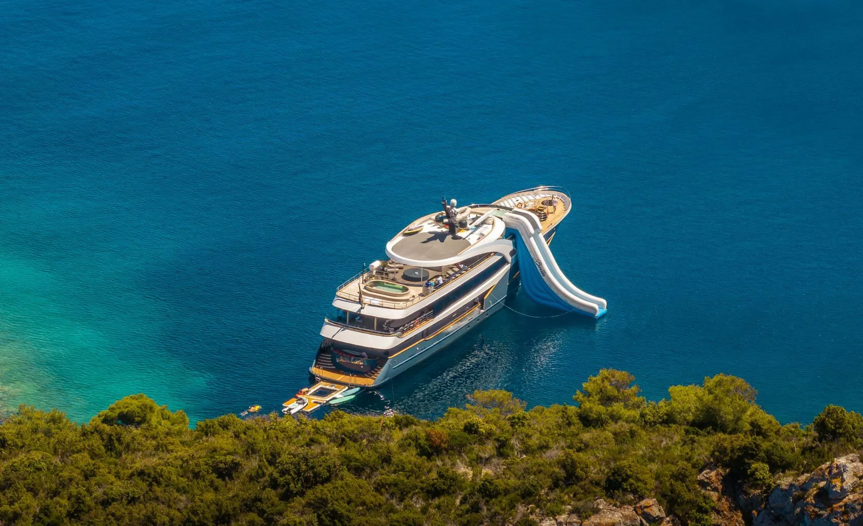 A large luxury yacht with a water slide anchored near a green, tree-covered shoreline in clear blue water. The yacht has multiple decks and sunbathing areas visible.