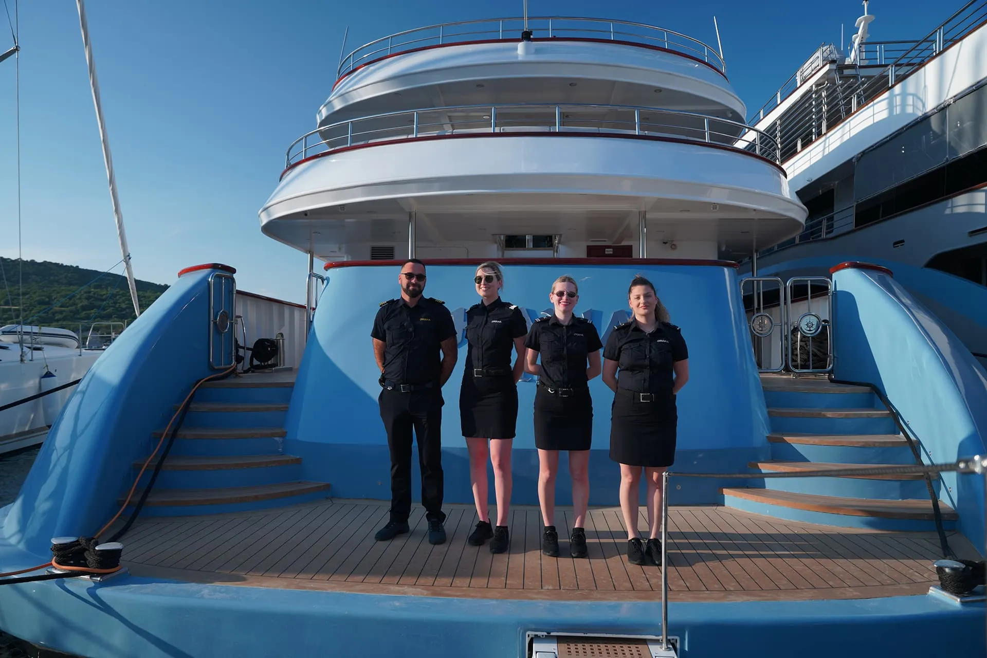 Four uniformed crew members stand on the deck of a blue and white yacht, posing for a photo with the back of the boat and a clear sky in the background.