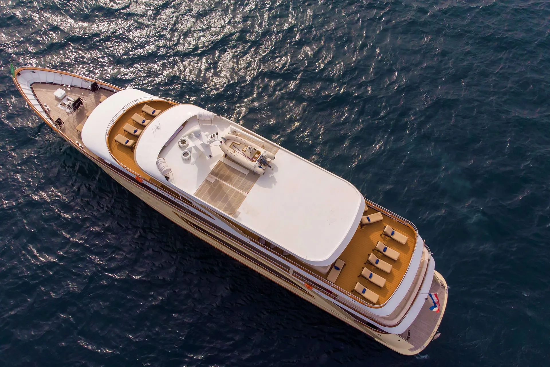 Aerial view of a luxurious yacht with a spacious deck, lounge chairs, and shaded areas, cruising on deep blue water under bright sunlight.