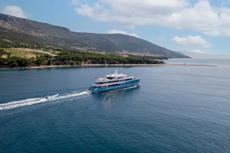 A large blue and white yacht sails near a green coastline, followed by a small speedboat on calm blue water. Hills and a partly cloudy sky are visible in the background.