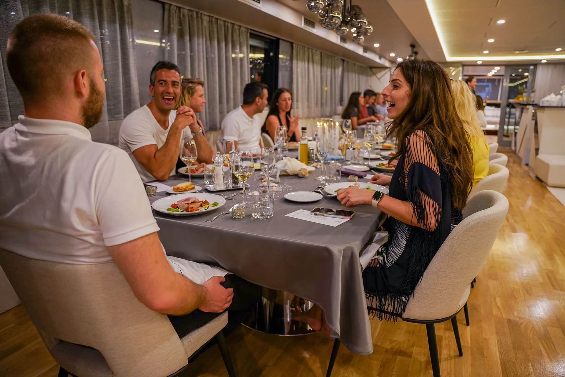 A group of people are sitting around a dining table, enjoying food and conversation in a modern, well-lit room with wooden floors and large windows. Plates, glasses, and candles are on the table.