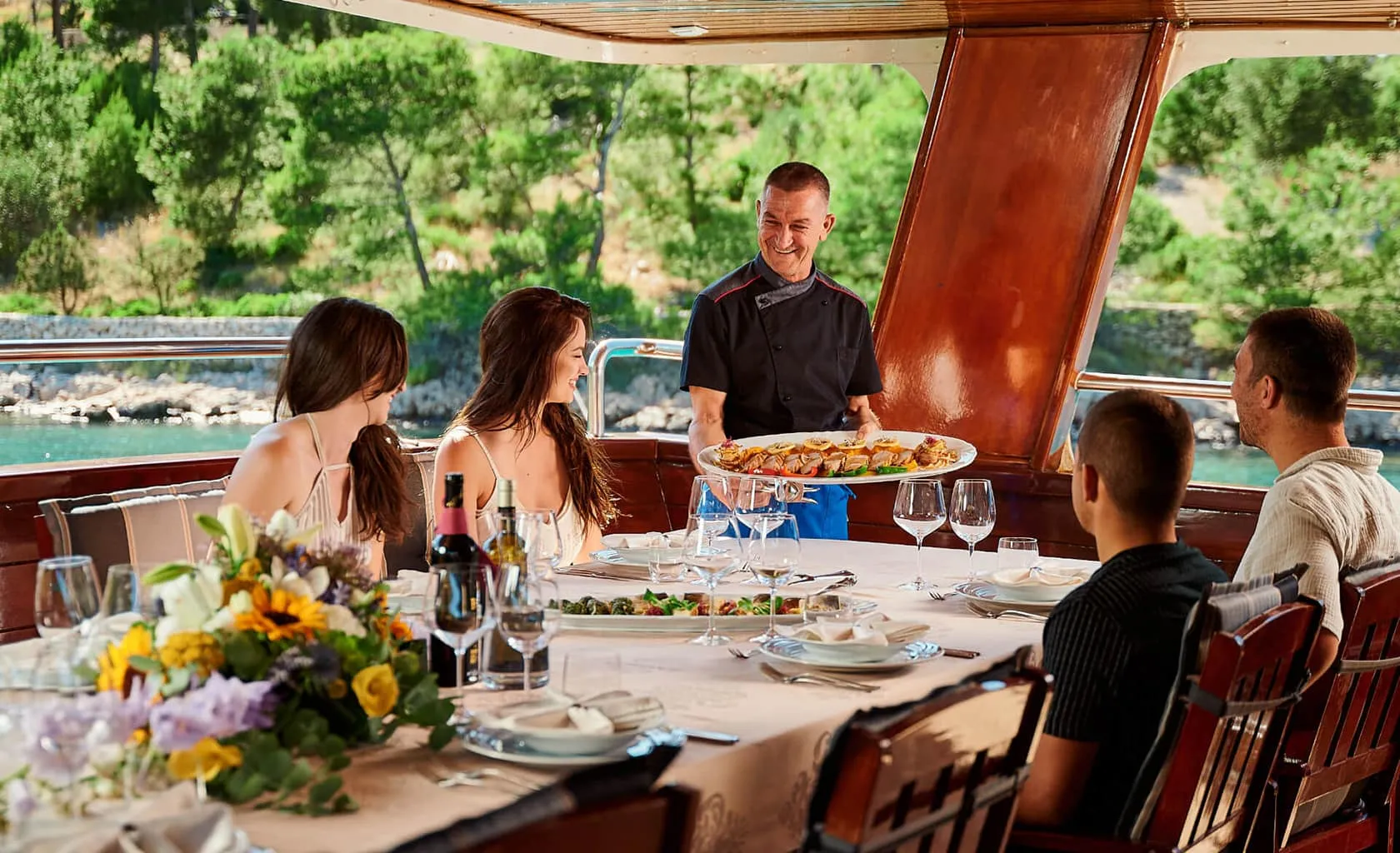 A chef serves a large platter of food to three women and one man seated at a dining table on a boat, surrounded by flowers, wine, and plates, with a view of trees and water in the background.