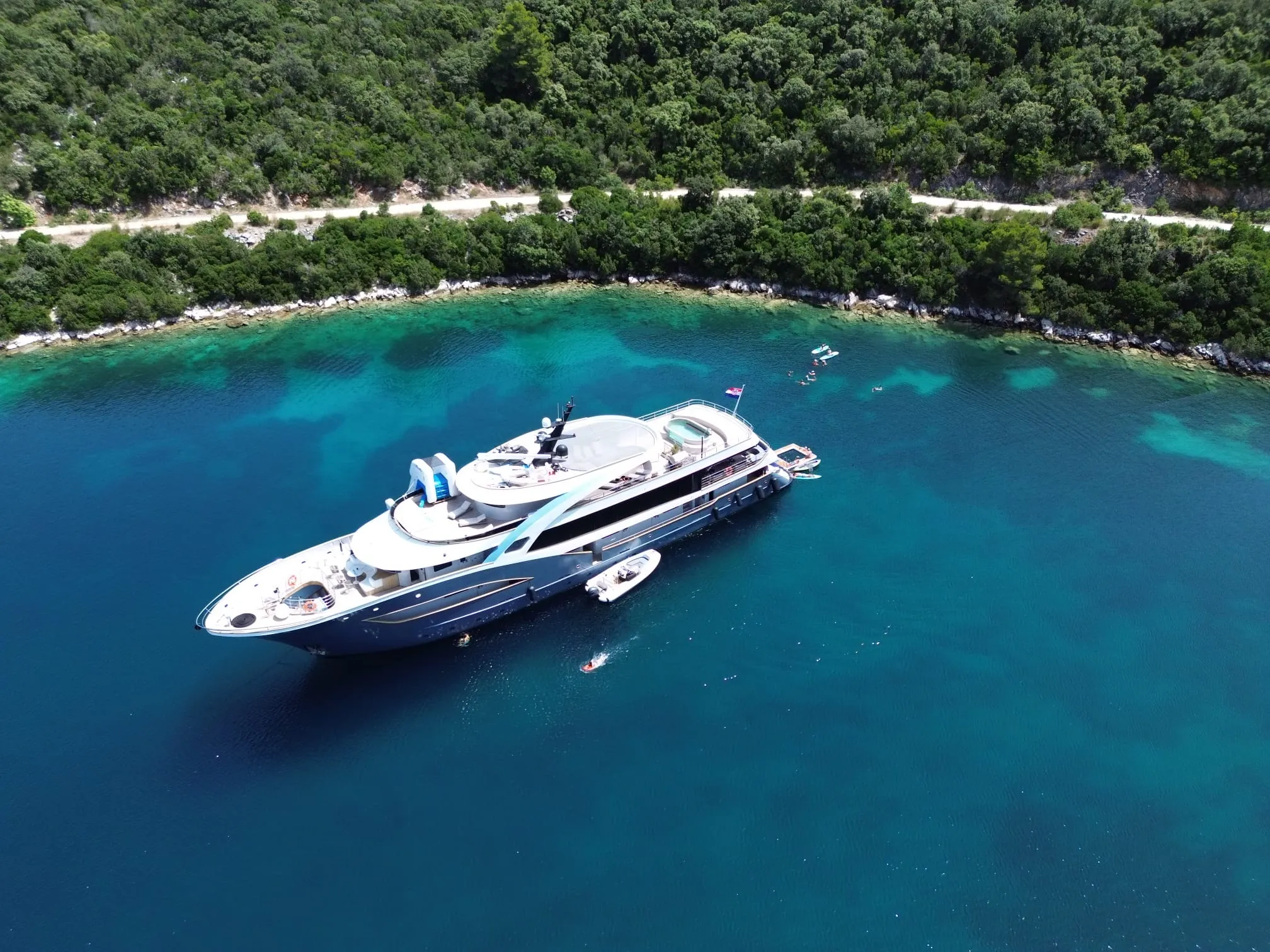 A large luxury yacht anchored in clear turquoise water near a lush, green coastline, with smaller boats floating nearby and a winding road visible through the trees.