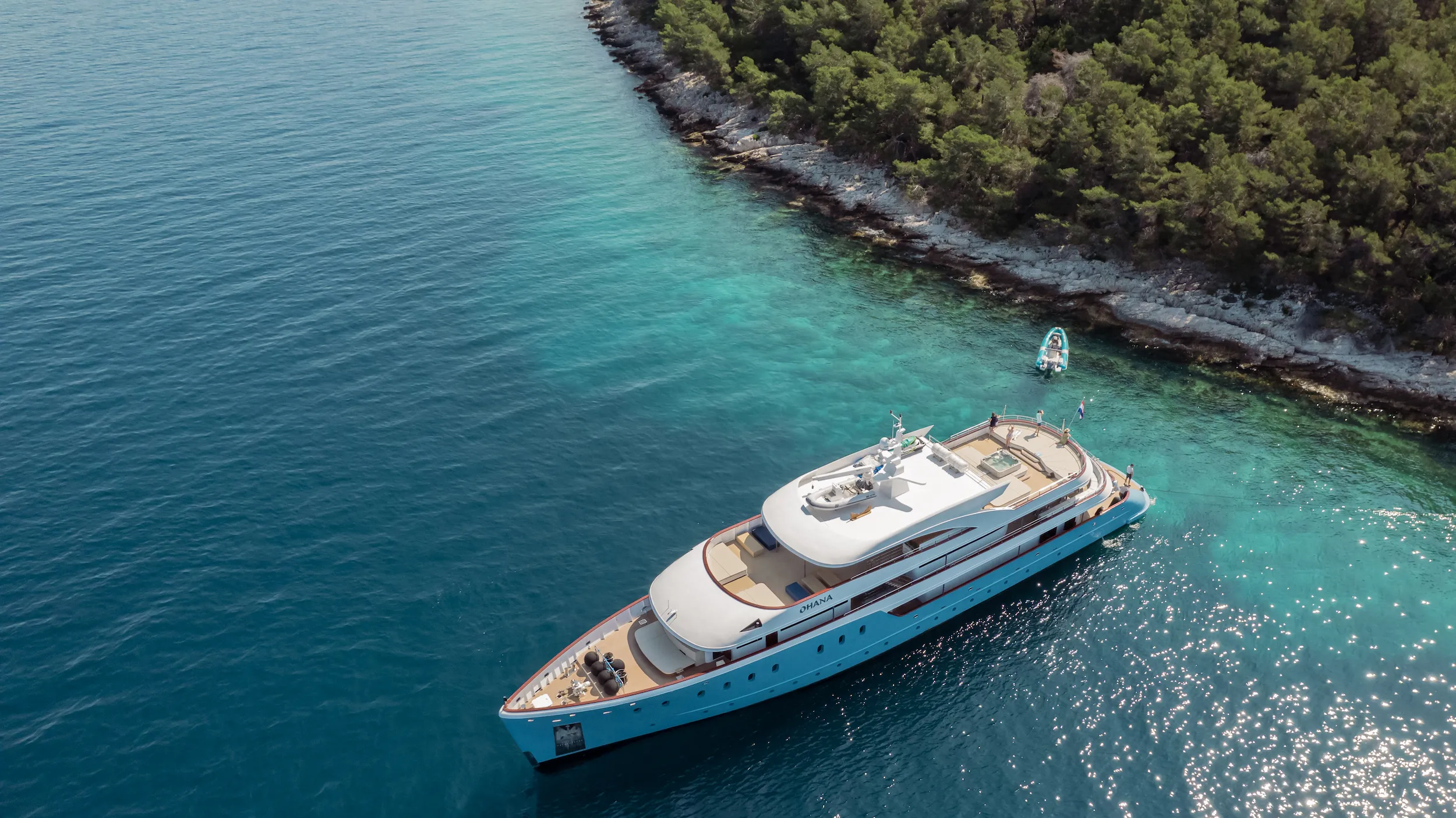 A large white yacht floats on clear blue water near a forested coastline, with a small dinghy nearby and dense green trees lining the rocky shore.