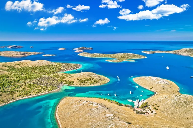 Aerial view of small, green islands surrounded by clear turquoise and blue sea under a bright sky with scattered clouds. Several boats are sailing in the calm water between the islands.
