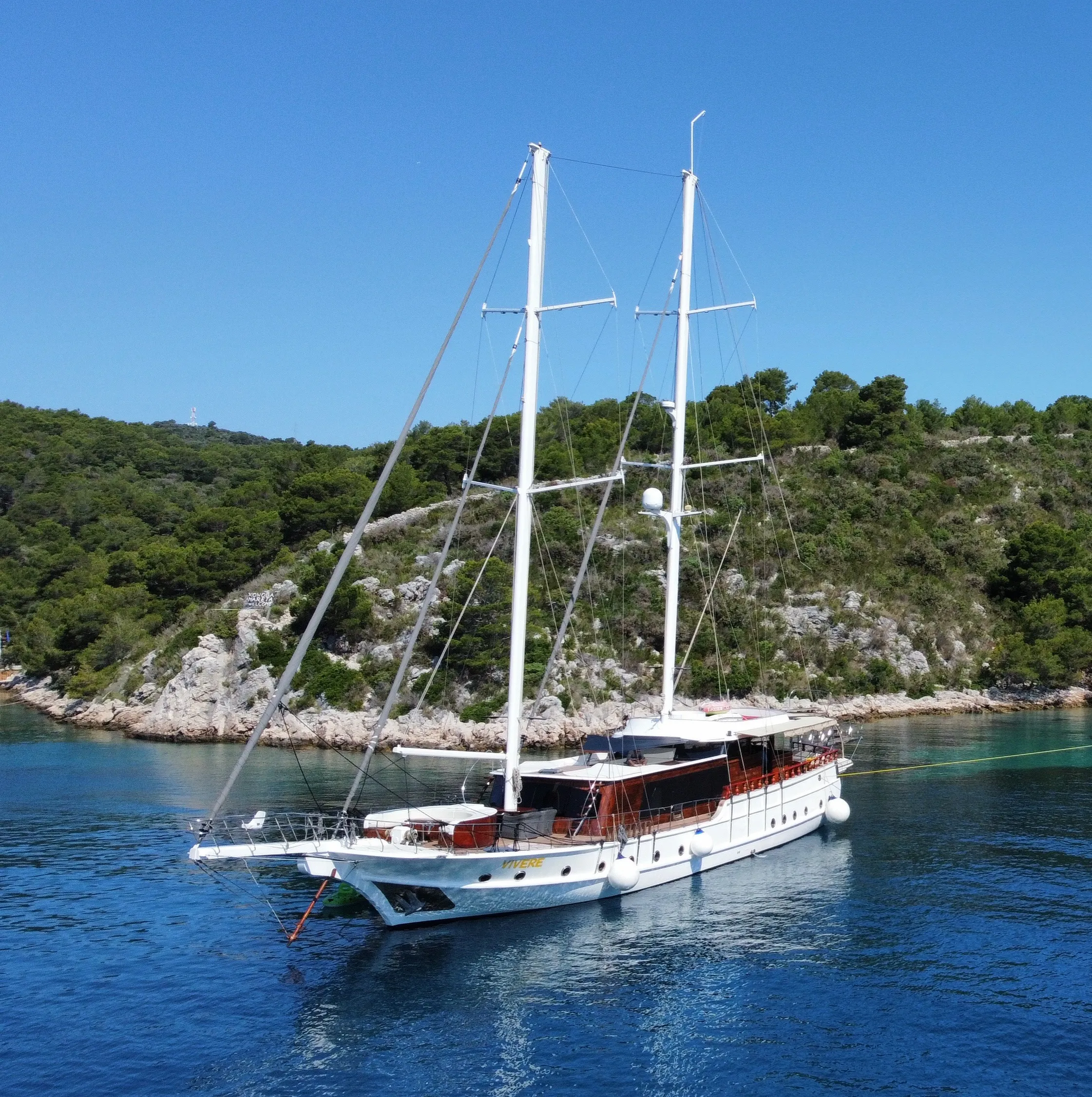 A white sailing yacht with two tall masts is anchored in calm blue water near a forested, rocky shoreline under a clear blue sky.