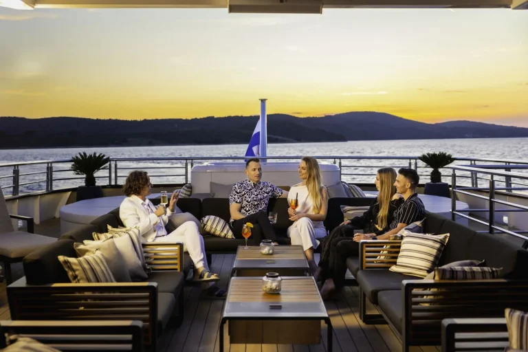 Five people sit and chat on a yacht deck at sunset, holding drinks. The group is surrounded by cushions and outdoor furniture, with calm water and hills visible in the background.