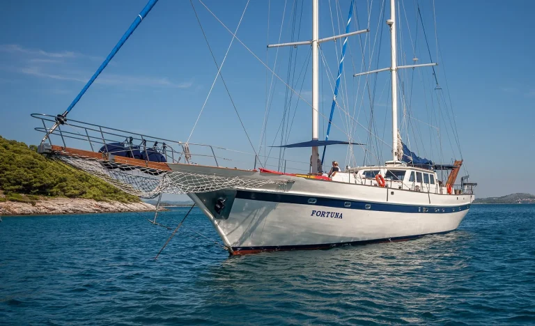 A white sailboat named “Fortuna” anchored on calm blue water near a rocky, tree-lined shore under a clear sky. The boat has two tall masts and blue accents.