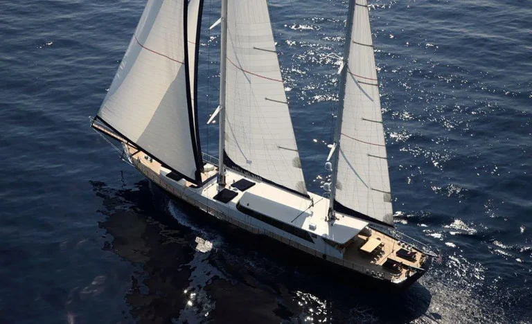 Aerial view of a large sailing yacht with two masts gliding on calm, dark blue water. Sunlight reflects off the sails and the sea, creating a bright, peaceful atmosphere.