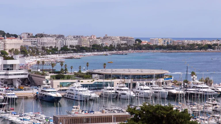 A marina filled with yachts and boats sits beside a curved, modern building on a sunny coastline, with sandy beaches, palm trees, and white buildings stretching along the waterfront.