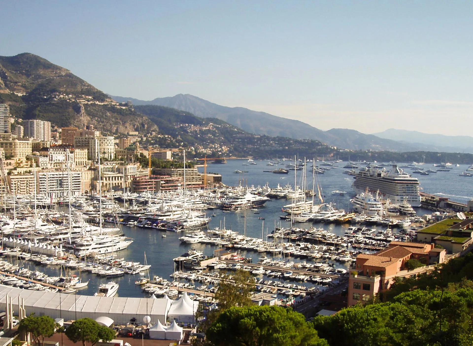 A coastal city with a busy marina full of yachts and boats, surrounded by tall buildings and mountains in the background under a clear sky.