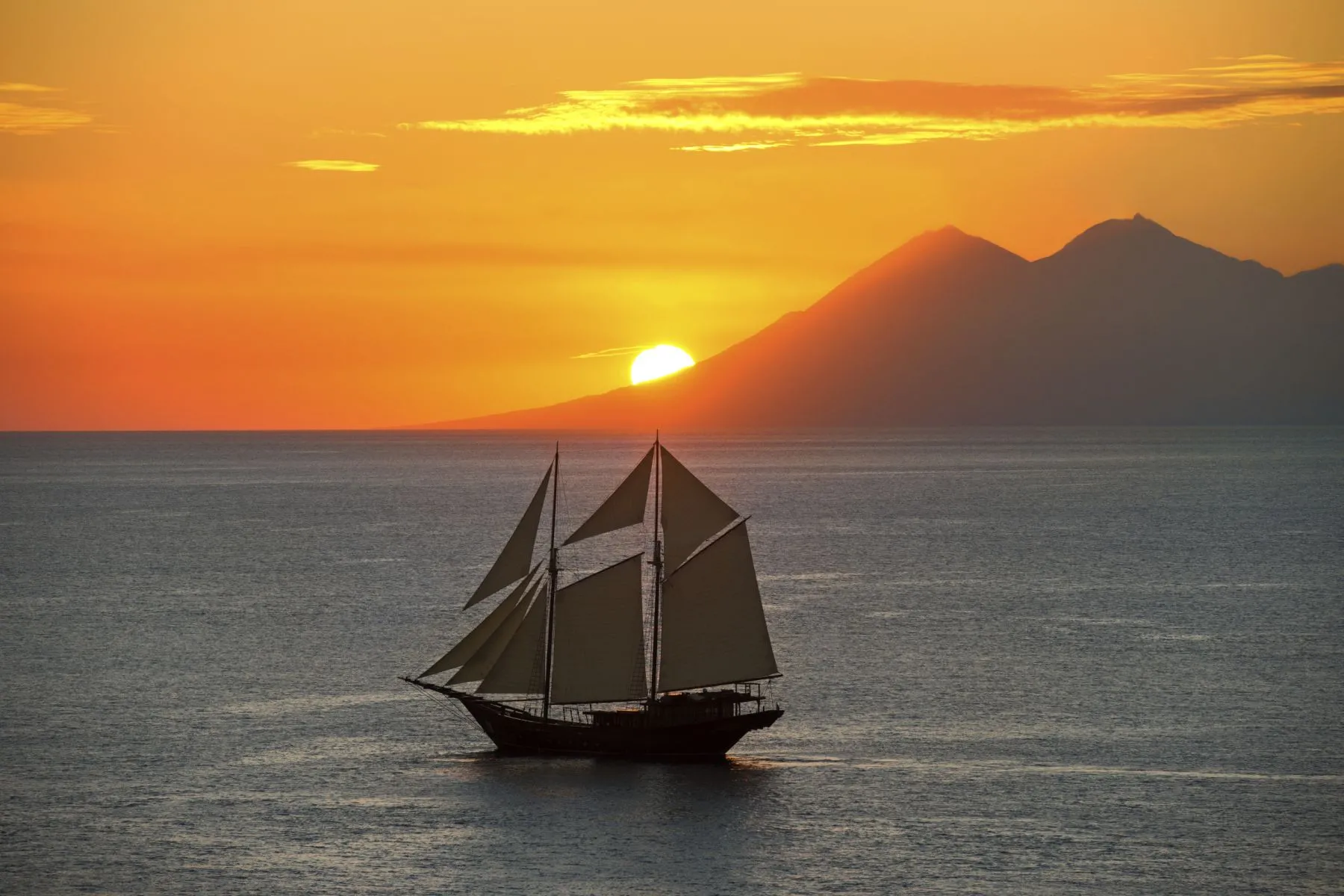 A sailboat with raised sails glides across calm water at sunset, with the sun partially hidden behind distant mountains and an orange sky reflected on the sea.