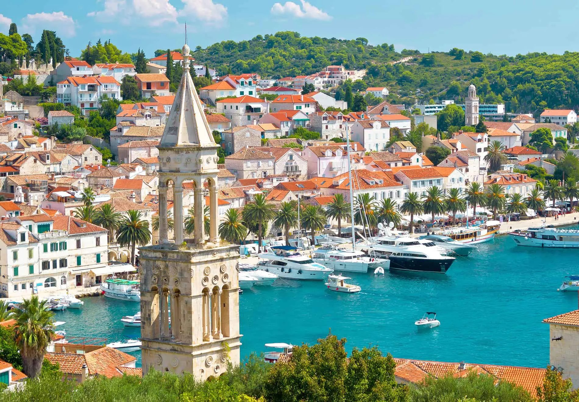 A scenic coastal town with red-roofed buildings, a stone bell tower in the foreground, palm trees along the waterfront, and yachts docked in the turquoise harbor, nestled against lush green hills.