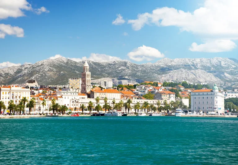 A scenic coastal city with historic buildings, red-tiled roofs, palm trees, and a prominent bell tower, set against a backdrop of mountains under a blue sky with scattered clouds. Calm blue water is in the foreground.