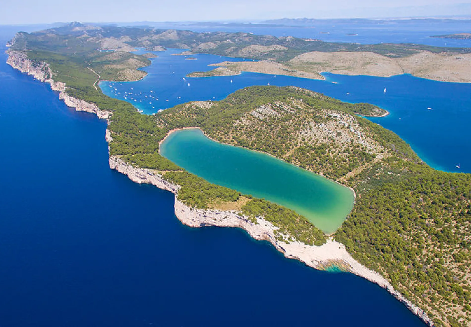 Aerial view of a rugged coastline with turquoise and deep blue waters, lush green islands, and a prominent saltwater lake separated from the sea by a narrow strip of land.