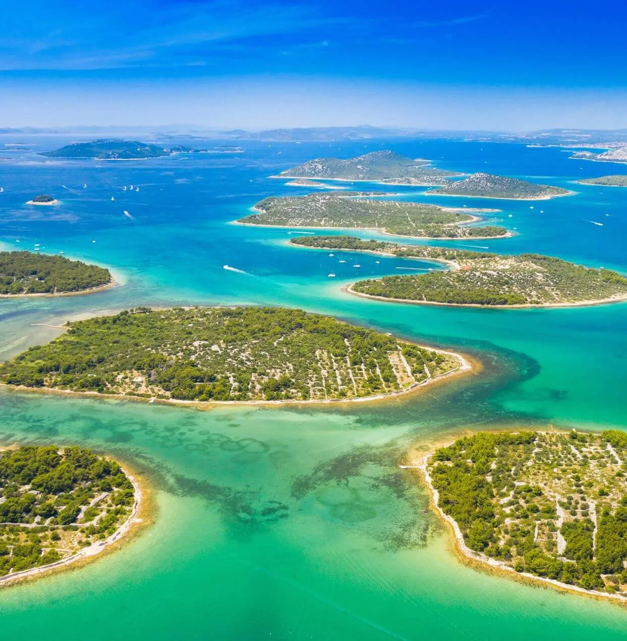 Aerial view of lush green islands surrounded by clear turquoise water, with a few boats scattered across the sea under a bright blue sky.