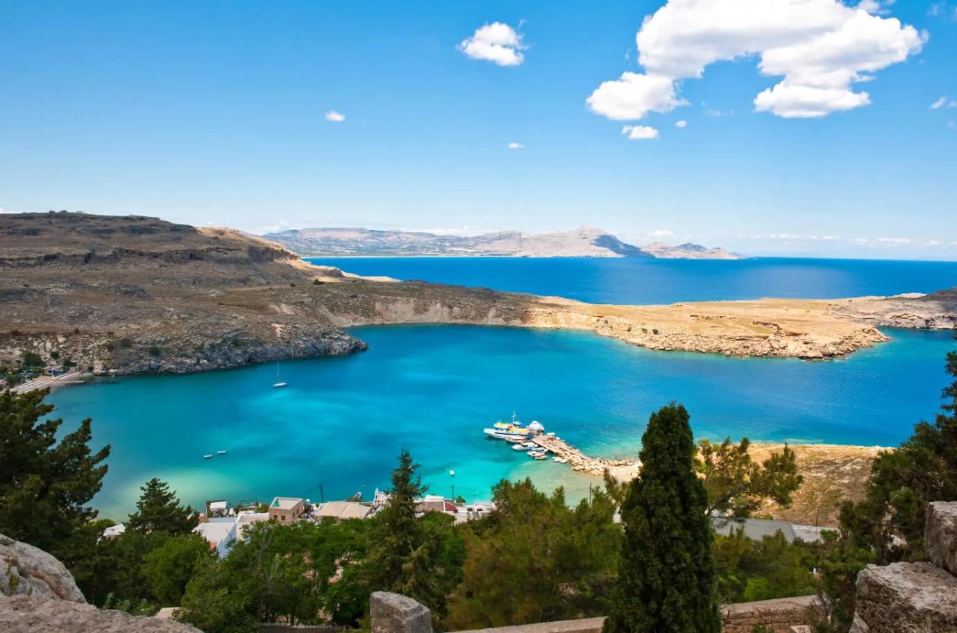 A scenic view of a turquoise bay surrounded by rocky hills and greenery, with boats docked at a small pier. The clear blue sky is dotted with a few clouds, and distant mountains rise on the horizon.