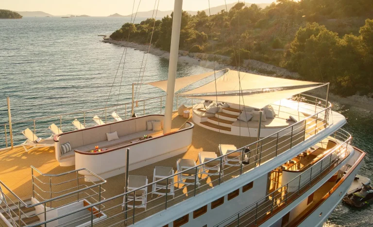 A luxury yacht deck with lounge chairs, a shaded seating area, and a view of the coastline and sea at sunset. The yacht is anchored near a wooded shore with calm, blue water.