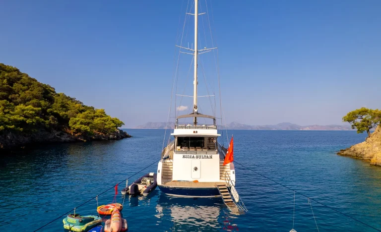A white yacht named Kasla Sultan is anchored in a calm, blue bay surrounded by green, tree-covered hills under a clear sky. Several small boats and floatation devices are tied to the yacht.