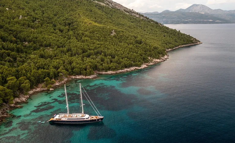 A sailboat is anchored in clear turquoise water near a tree-covered coastline, with hills and mountains visible in the background under a partly cloudy sky.
