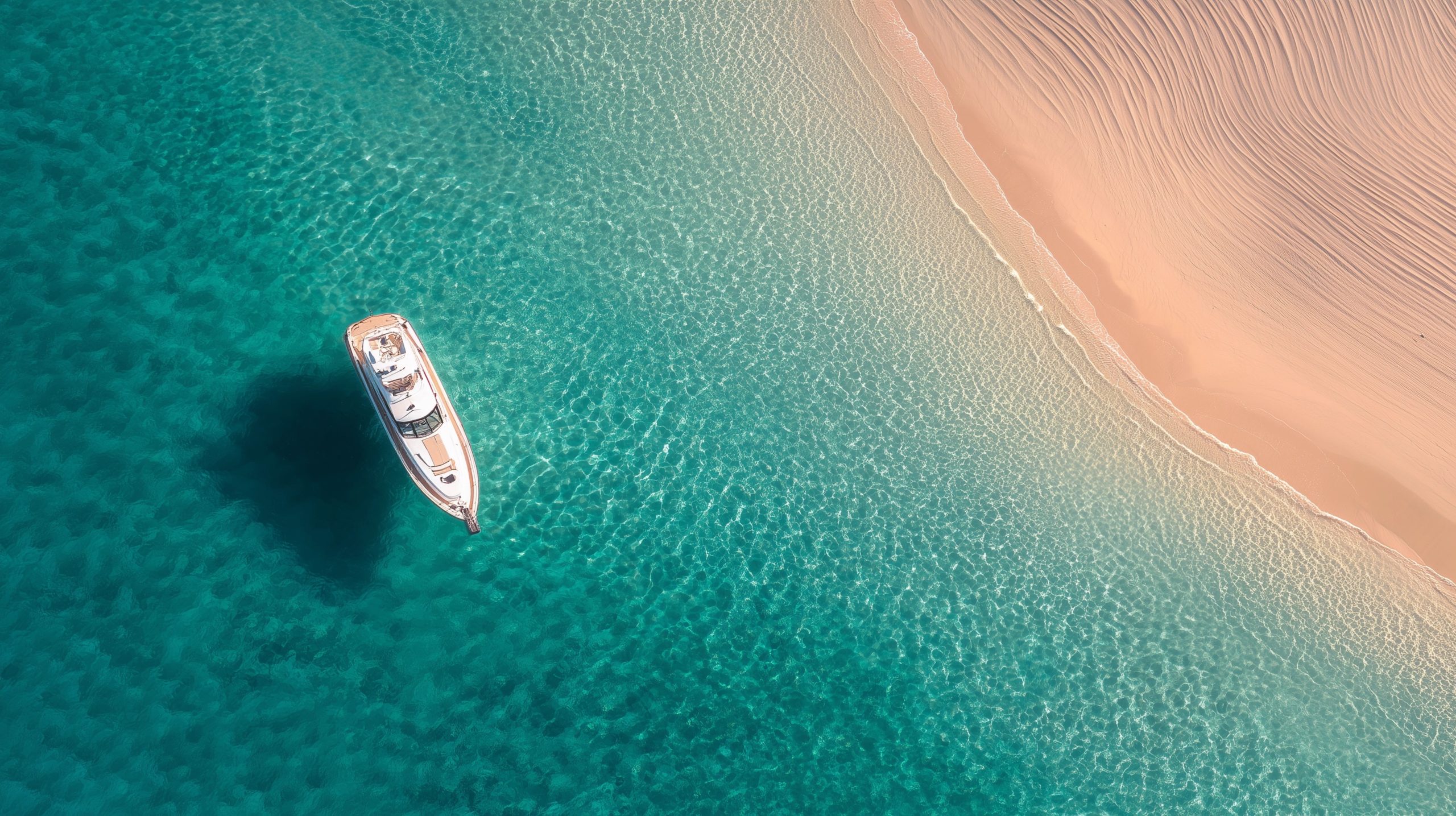 A white yacht floats on clear turquoise water near a sandy beach with gentle ripples, viewed from above. The beach curves along the right side, and the boat casts a shadow on the water.