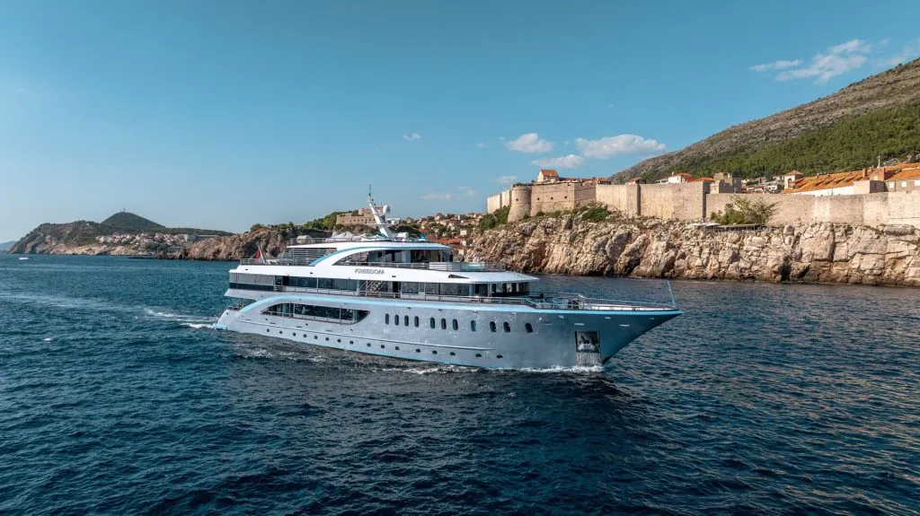 A large, modern yacht rental sails on blue water near a rocky coastline with historic stone buildings and a mountainous backdrop under a clear sky.