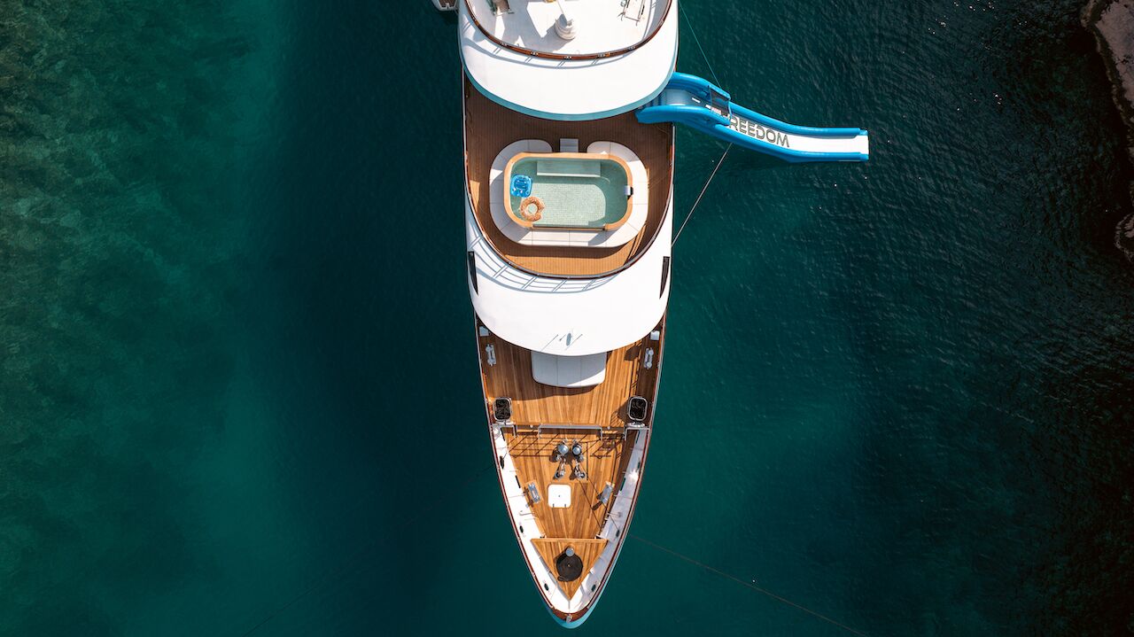 Aerial view of a luxury yacht charter anchored in clear blue water, with a person relaxing in a small pool on the deck and a blue slide extending from the upper deck into the sea.