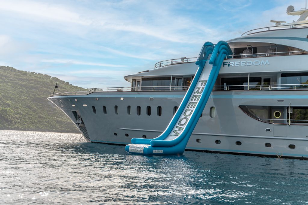 A luxury yacht for charter anchored near a lush, green coastline features a large blue inflatable slide labeled FREEDOM on its upper deck, extending down to the water.