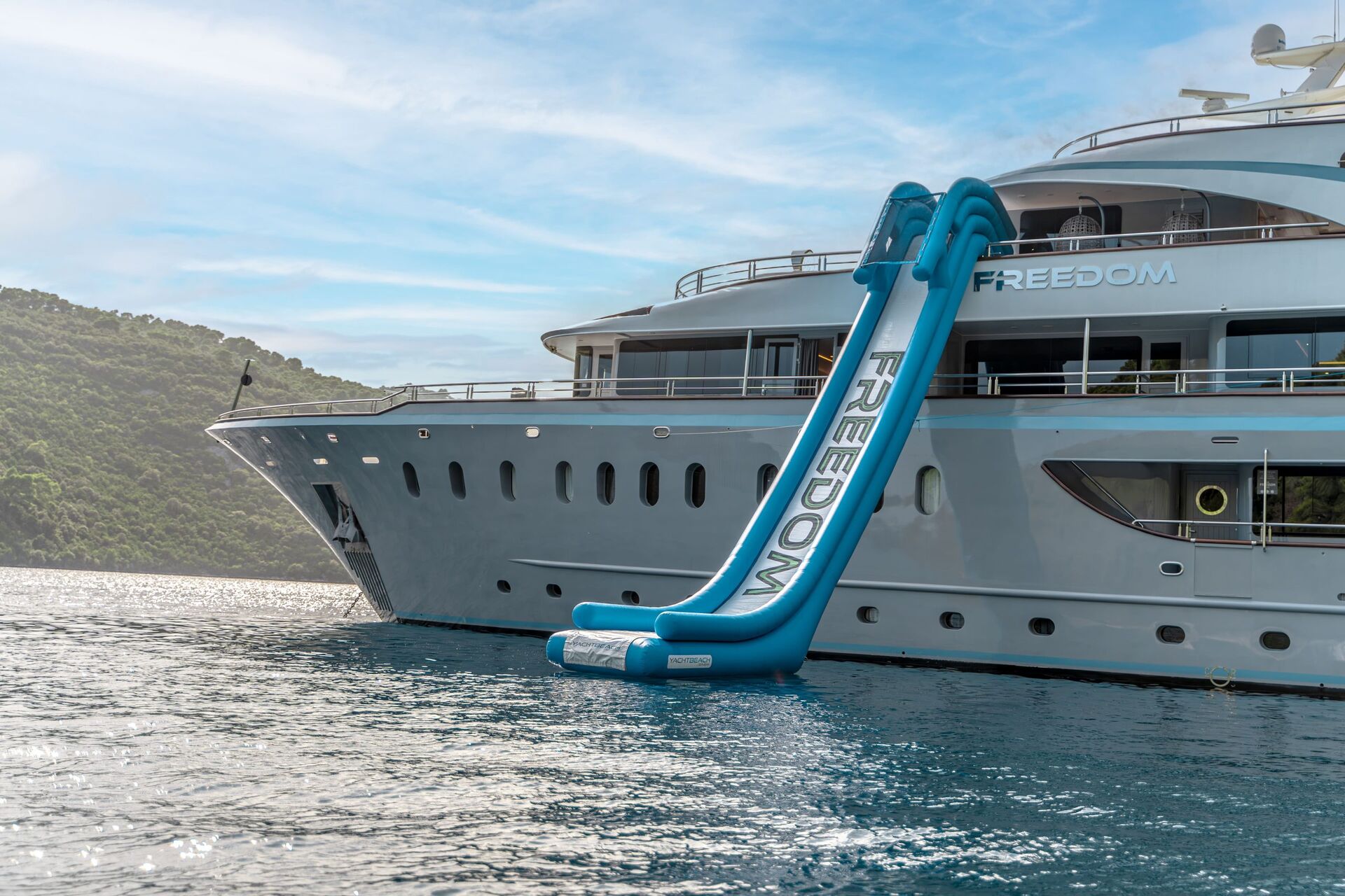 A luxury yacht for charter anchored near a lush, green coastline features a large blue inflatable slide labeled FREEDOM on its upper deck, extending down to the water.
