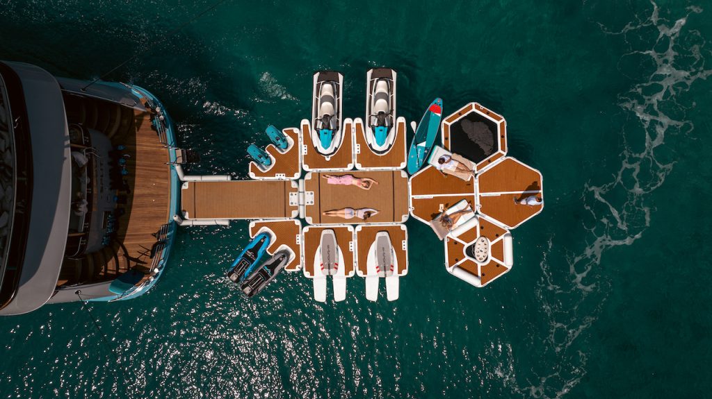 Aerial view of people relaxing on a floating dock with paddleboards, jet skis, and modular platforms connected to a yacht rental on clear turquoise water, perfect for those seeking luxury yachts for charter.