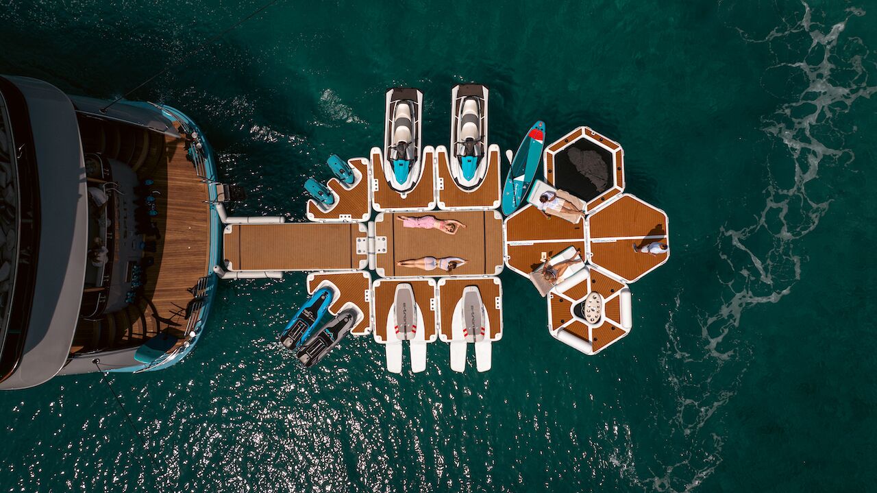 Aerial view of people relaxing on a floating dock with paddleboards, jet skis, and modular platforms connected to a yacht rental on clear turquoise water, perfect for those seeking luxury yachts for charter.