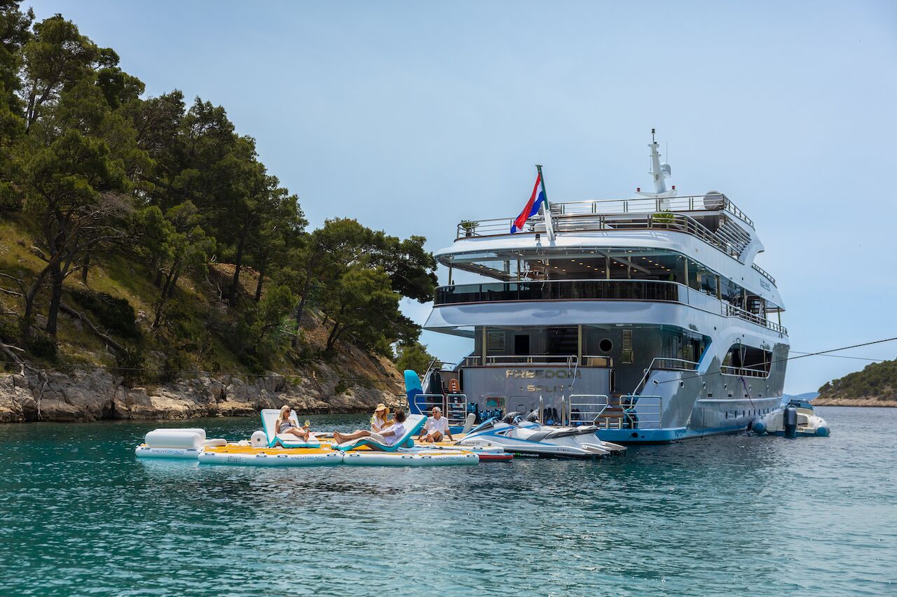 A large luxury yacht charter is anchored near a forested shoreline. Several people relax on inflatable rafts in the calm blue water beside the yacht under a clear sky.