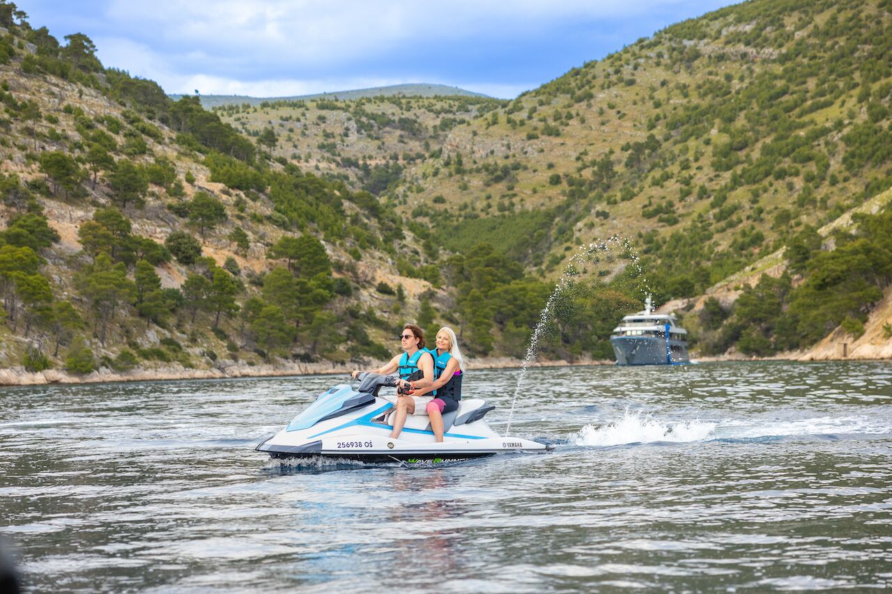 Two people wearing life jackets ride a jet ski on a calm river surrounded by green hills. In the background, a large boat available for yacht charter is visible on the water under a partly cloudy sky.