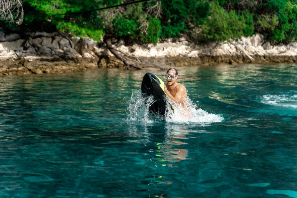 A person wearing goggles rides a motorized surfboard on clear turquoise water near a rocky, tree-lined shoreline, creating splashes as they move—an exciting scene perfect for those enjoying a yacht charter adventure.
