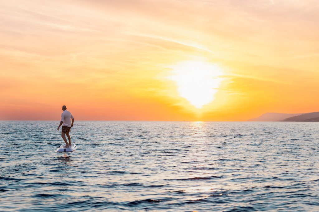 A person rides an electric surfboard over calm ocean water at sunset, with the sky glowing orange and yellow and distant hills on the horizon—an idyllic scene perfect for those enjoying a yacht charter nearby.