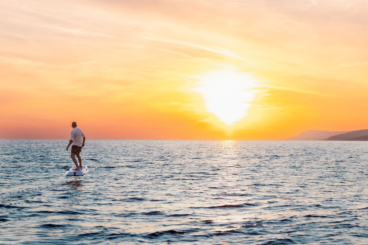 A person rides an electric surfboard over calm ocean water at sunset, with the sky glowing orange and yellow and distant hills on the horizon—an idyllic scene perfect for those enjoying a yacht charter nearby.