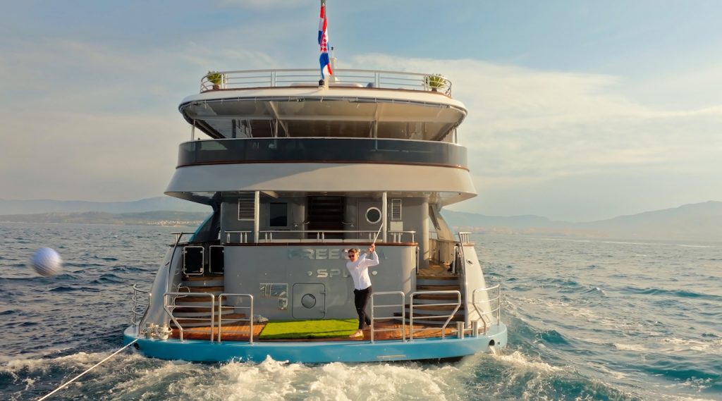 A person stands on the back deck of a large yacht rental, raising their arms as the boat glides through calm blue water. The sky is partly cloudy, and distant mountains are visible on the horizon.