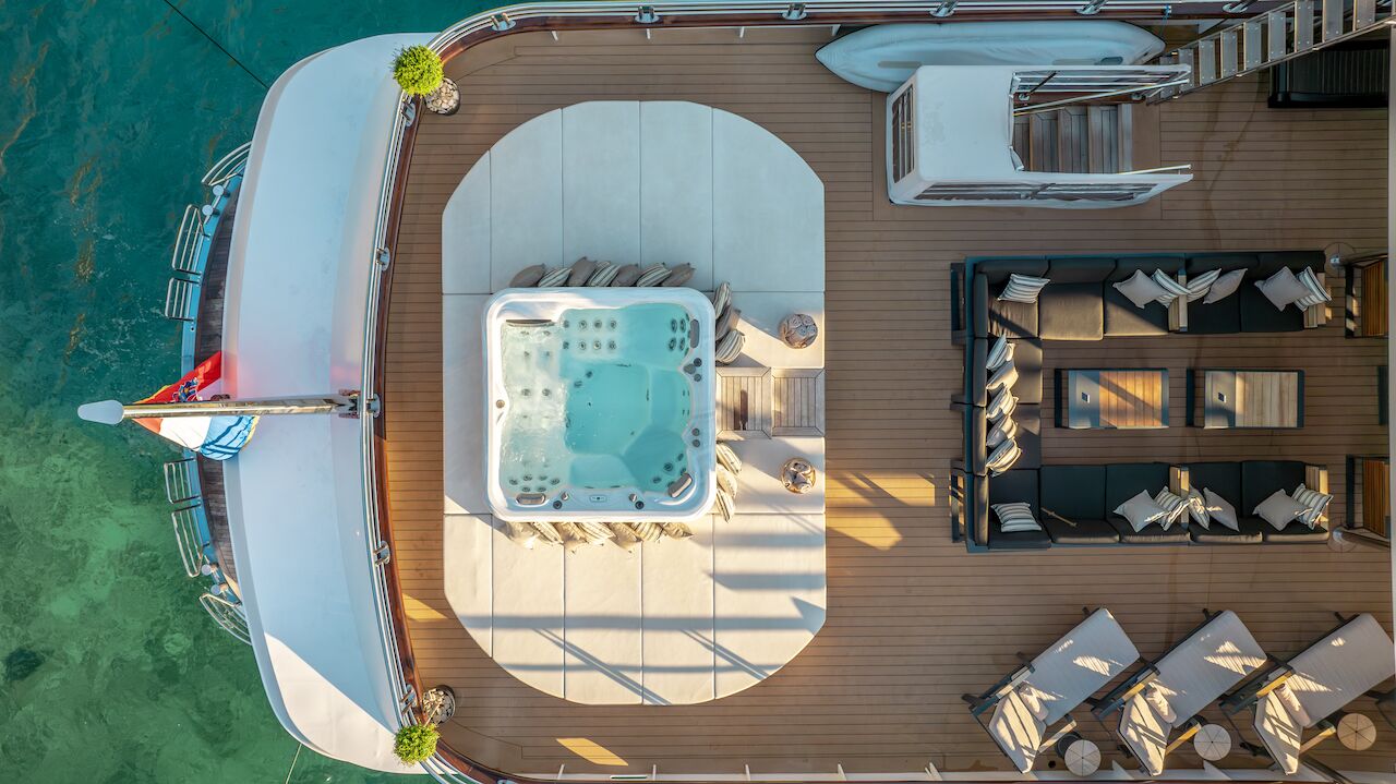 Aerial view of a yacht rental deck featuring a hot tub at the center, surrounded by lounge chairs, sofas, tables, and potted plants, with clear turquoise water visible beside the yacht.