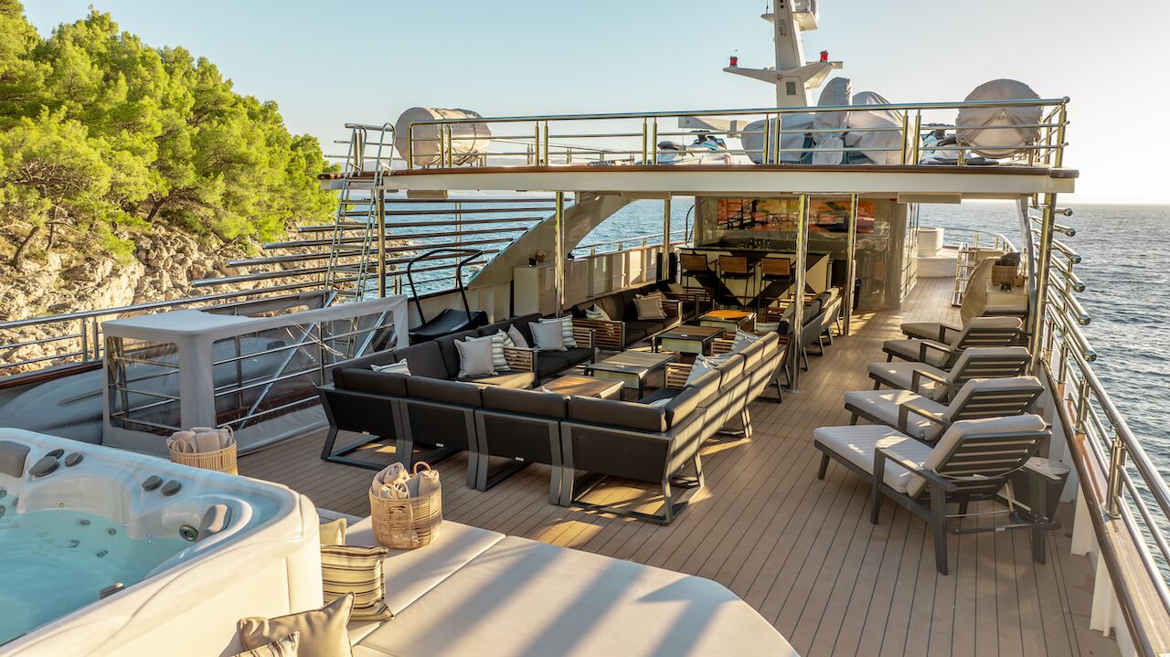 Spacious yacht deck with lounge chairs, sofas, tables, and a hot tub, overlooking the sea; ideal for a relaxing yacht charter. Trees and rocky shore appear on the left, as sunlight illuminates the inviting scene.