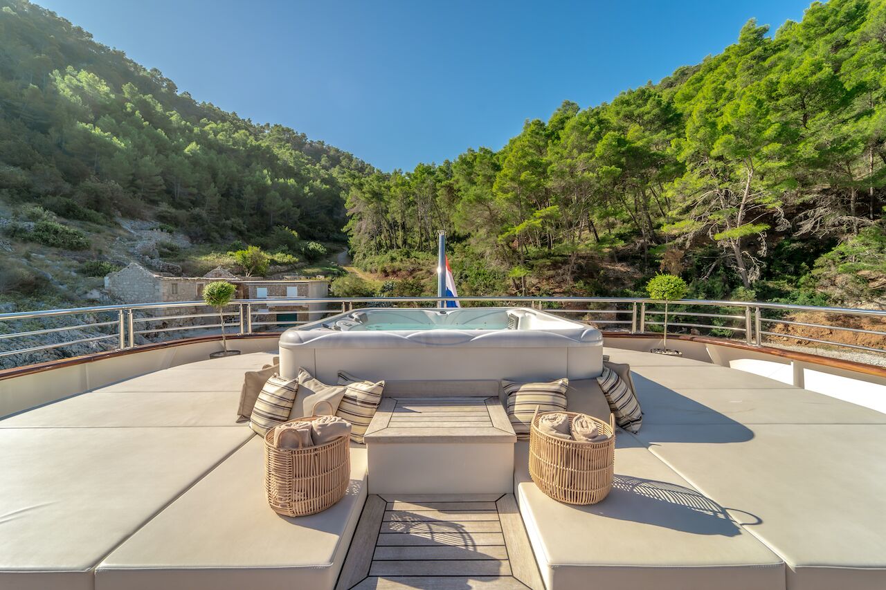 A luxurious yacht charter deck with beige cushioned seating, wicker ottomans, and a hot tub overlooks lush green hills under a clear blue sky.
