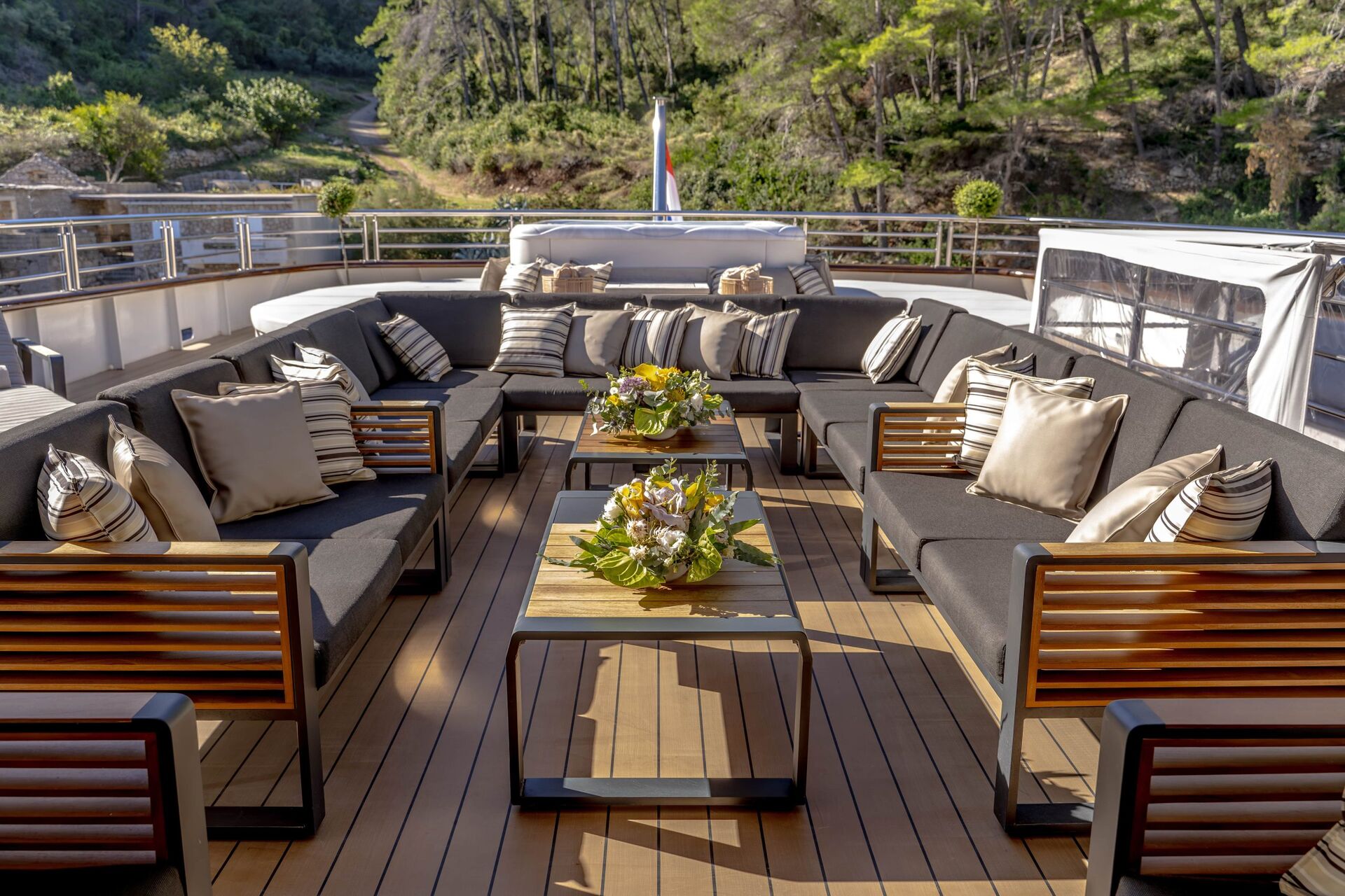 Outdoor yacht deck with wooden flooring, dark cushioned seating, striped pillows, and three tables with floral centerpieces—perfect for a relaxed yacht charter experience. Surrounded by greenery and trees in the background.