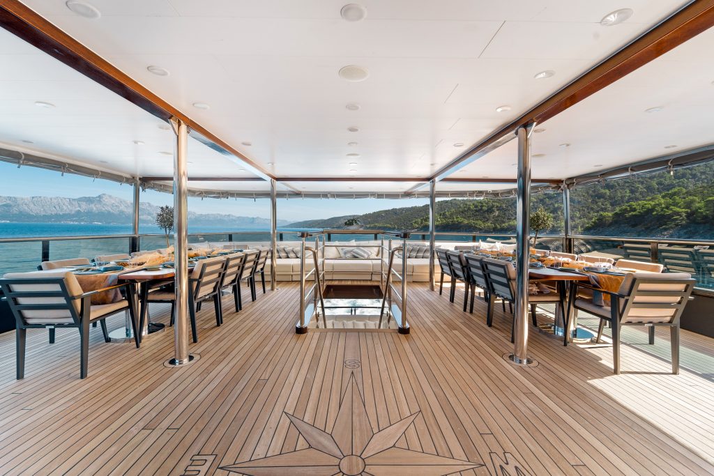Spacious yacht deck with wooden flooring, dining tables, and chairs arranged on both sides. Ideal for a memorable yacht charter, the deck overlooks scenic blue water, mountains, and lush greenery under a partly cloudy sky.