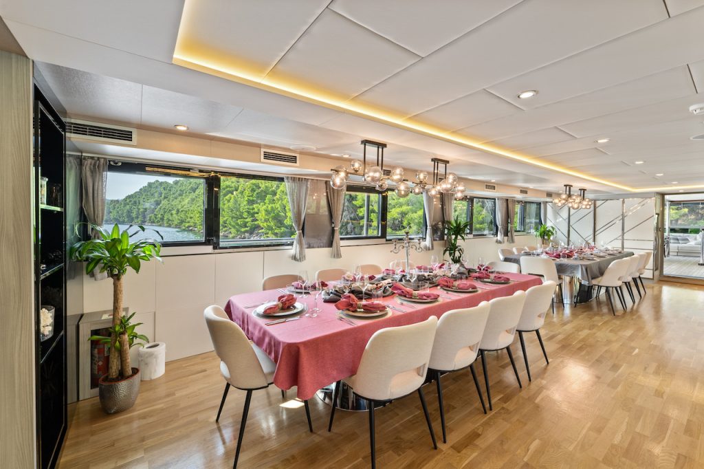 Elegant dining area on a yacht charter with two tables set with pink tablecloths, plates, and glasses. Large windows show green trees outside. Modern lighting and plants decorate the space with wood flooring.
